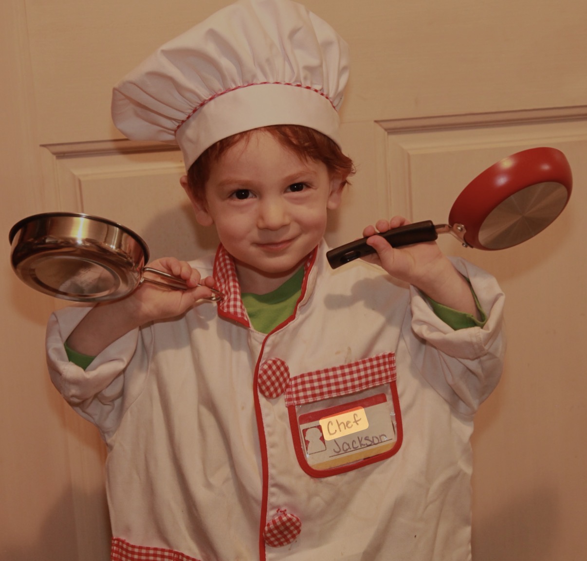 Three-year-old in a chef costume holding play pans, name tag reads Chef Jackson.